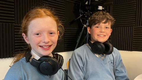 Caoimhe and James sit side by side in a recording studio, each wearing large over‑ear headphones around their necks. They are dressed in matching light‑blue school jumpers. Acoustic foam panels line the walls, and a camera tripod is set up in the background.