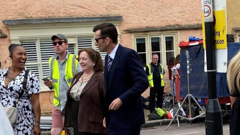 David Tennant in a suit and Claire Rushbrook surrounded by people in high-vis and filming equipment.