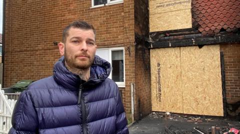 Dean Ray looking into the camera with a slight smile. He has brown, short hair which has been gelled to the side on top. He is wearing a blue puffer coat and has tattoos on his neck which are just visible. He is standing in front of the burnt house which has cardboard covering the door and window above it. There is rubble on the floor.
