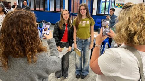 In the foreground are two mothers, standing in front of their daughters, taking a picture of them together holding their GCSE exam results. The two daughters in the background are wearing casual clothes and smiling.