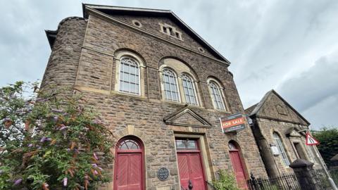 Front façade of the chapel with three double wooden doors side by side painted dark red and four arched windows above. An orange for sale sign has been attached to the wall at the front.