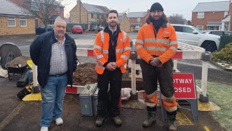 Ozzy O'Shea on the left in a white shirt and navy jacket. He is stood next to two Cadent engineers who are in orange high-vis jackets.