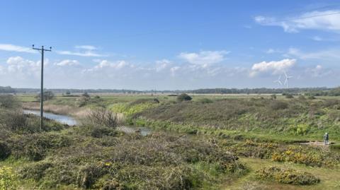 A general view of the Benacre sluice. A large green field can be seen with a river running through the middle of it. A pylon can be seen in the distance as well as two wind turbines.