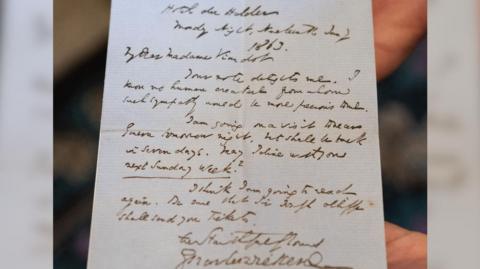 An unseen curator holds up a letter handwritten by the author Charles Dickens.