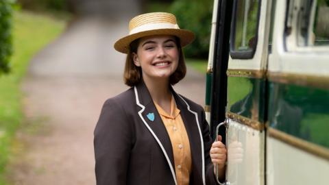 Darrell stands smiling by a bus wearing her Malory Towers uniform