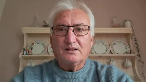 Peter Horgan has short white hair and glasses. He is wearing light blue sweater. Behind him is a cream‑coloured shelving unit filled with decorative plates