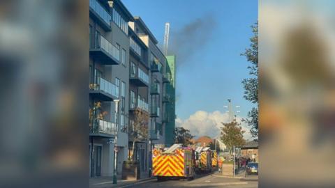 Three fire engines stopped outside a modern-looking block of flats, with glass balconies. There's smoke rising from the roof. It's a sunny day and the sky is blue.