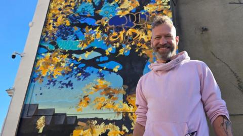 Joe Webster stood to the right of the large mural on the side of the building. He is wearing a pink hooded jumper, and is looking down at the camera while smiling. In the background is the blue and yellow tree with graffitied words amongst the foliage. 