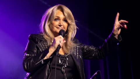 Bonnie Tyler is wearing a jacket and a black t shirt, while holding a microphone and singing.