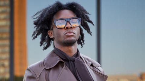 D4vd is pictured in large, black-rimmed glasses and a brown leather coat