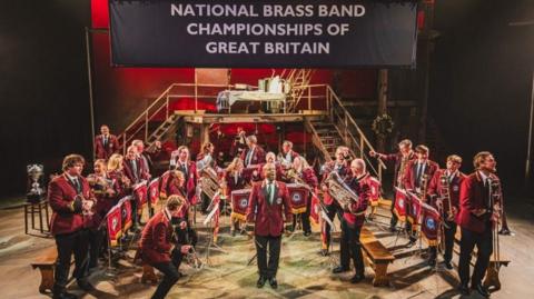 A brass band on a stage with their instruments - they are dressed in red blazers and black trousers and there is a sign which reads 'National Brass Band Championships of Great Britain'