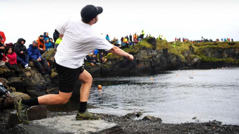 A man in a black cap and white t shirt throws a stone out on to water as a crowd watches on a rocky bank behind him on a grey cloudy day.