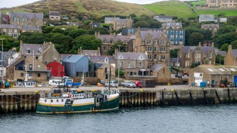 Stromness harbour with a green and yellow boat tied up and buildings and cars in the background