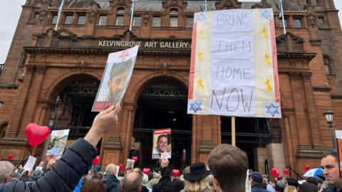 Crowds gathered outside Kelvingrove art gallery. They are holding banners, some show pictures of hostages another has 'bring them home now' written on it.