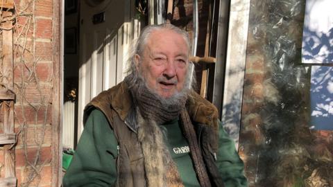 Richard Owen Vinton wearing a brown scarf, gilet-type jacke with a green top underneath, and standing outside his front door