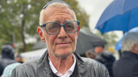 Mark Goldstone, who has white hair, glasses, a yarmulke skullcap and a black jacket, looks into the camera. He is drenched with rain water and people can be seen in the background holding umbrellas. 