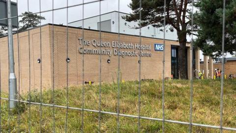 A large yellow bricked building with "The Queen Elizabeth Community Diagnostic Centre" written in silver letters on the front. It's photographed through a fence to show work is still underway and it's not quite open yet. 