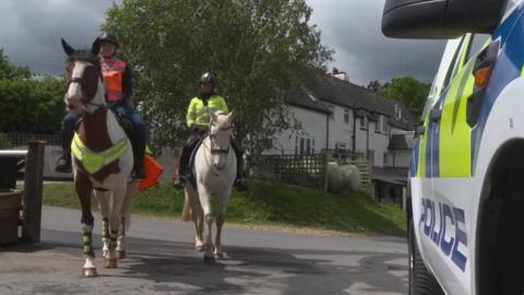 Two horses are seen approaching the camera with riders wearing high-viz jackets and a police car seen to the right. 