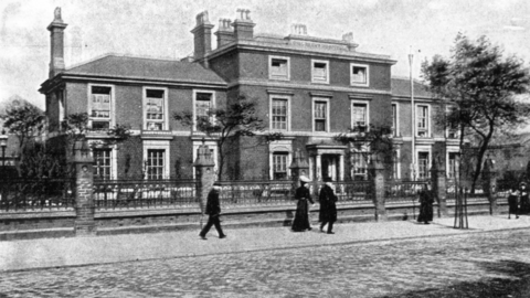 Black-and-white historical photograph of a large, two-story brick hospital building with multiple chimneys and tall sash windows, identified as “The Children’s Hospital, Birmingham.” A decorative iron fence runs along the front, separating the building from a cobbled street where several pedestrians in long coats and hats are walking. Sparse trees stand along the sidewalk, and the image has a slightly faded, archival appearance.