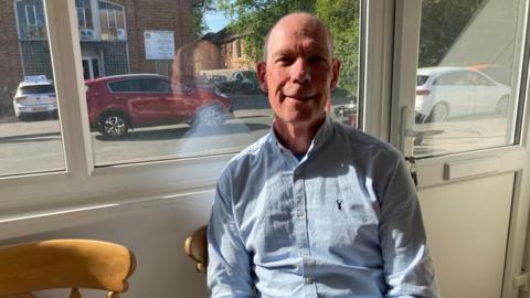 A man is sitting in a cafe with his back to the window. He is wearing a blue shirt and smiling at the camera