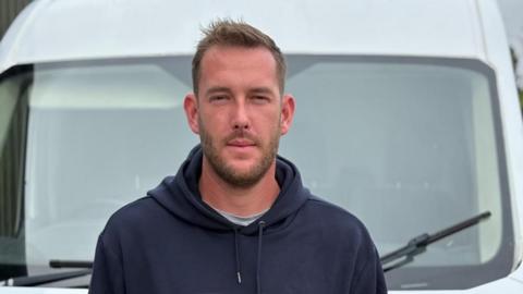 Toby Kennedy-Hall stares into the camera as he stands in front of his van. He wears a dark blue hoodie, with a grey T-shirt just visible underneath.