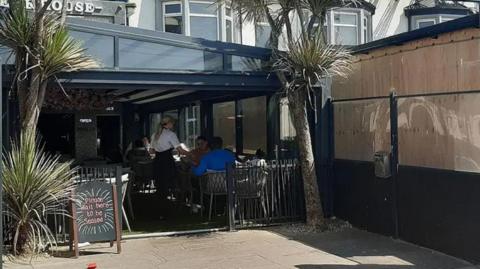 Street view of the restaurant showing a glass extension between the pavement and the restaurant. People are sitting in the extension while a waitress in a white shirt and with blond hair in a ponytail serves them. There is an A-board outside that says 'please wait to be seated'