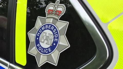 The side of a Humberside Police vehicle with the silver star crown logo with Humberside Police written in the middle. You can see bright yellow lines on the side of the car.