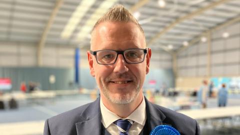 A man with short, grey spikey hair is pictured in a sports hall wearing glasses and a navy blue suit.