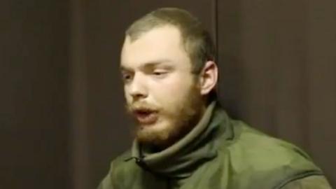 A man believed to be James Scott Rhys Anderson, 22, a former British Army soldier captured while fighting for Ukraine, is seen wearing green military clothing and sat in front of a plain brown background.