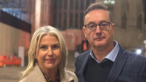 Christine wears a beige coat and has blonde hair, she stands next to Richard who wears a navy checked blazer and navy jumper with light blue shirt. He wear glasses and has short dark hair.
