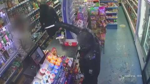 Teenager wearing a black hoodie points a knife at a shopkeeper who is standing behind the till.