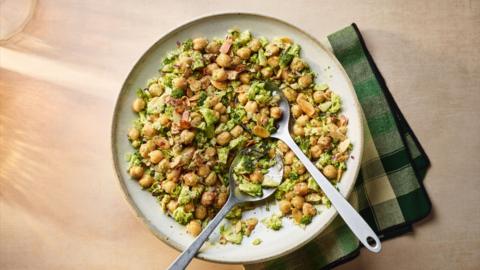 Aerial view of broccoli and chickpeas in a bowl with two spoons in it. It sits on a green checked napkin