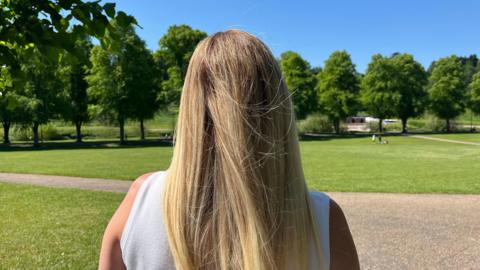 Adele pictured facing away from the camera. She's wearing a white vest and has her long blonde hair loose. She's stood in a park, with bright green grass, leafy trees, and a bright blue sky with strong sunshine. 