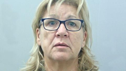 Stephanie White's mugshot against a plain pale blue/grey background. She has long wavy blonde hair tucked behind her ears, and navy blue framed glasses.