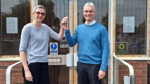 Two people standing outside, holding a set of keys between them. The woman, to the left has on a grey jumper, dark trousers and has short grey hair and glasses. The man has on a blue jumper, shirt and dark trousers. He also has glasses on and short grey hair. 