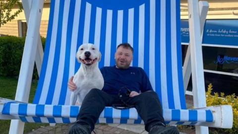 Paul Scott sits on a blue-and-white-striped over-sized deckchair with a cream-coloured dog sitting next to him. Mr Scott has short, dark hair and stubble, is wearing a blue jumper, and is smiling for the camera. 