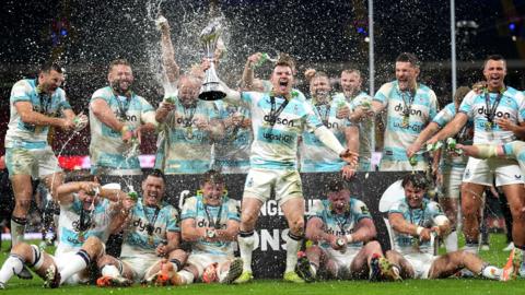 Bath players celebrate with the trophy and spray champagne in front of fans