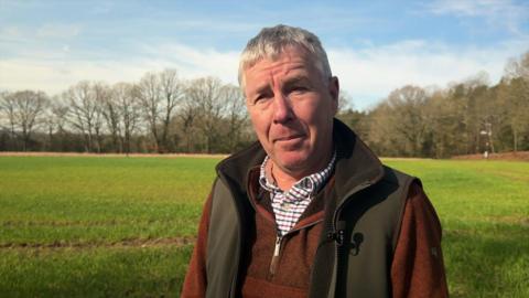 Peter Knight, a middle aged man with grey hair, wears a checked shirt, a brown jumper and a green gilet, as he stands in a field of wheat that is green and just beginning to grow.