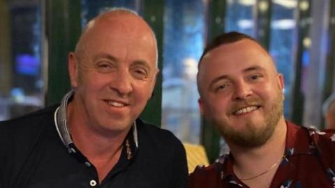 Two men, father and son smile at the camera. Peter, on the left, wears a black polo shirt, while Loughlin, on the right, has a beard, short light coloured hair and a maroon flowery shirt.  