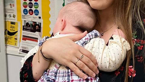 Generic image of a mother holding her baby girl in a room, in front of posters pinned up on a noticeboard at a family hub.