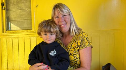 Jennifer Pattison is sitting inside a beach hut painted a similar yellow to her dress. She is holding her three-year-old grandson on her lap. He like her has blonde hair and he is wearing a black sweatshirt.