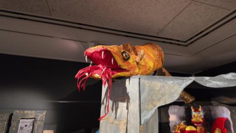 A yellow paper dragon placed on top of a shelf at the Story Museum. It's bright red mouth is open with red strands of paper hanging, representing blazing flames. Other dragon dolls could be seen underneath.