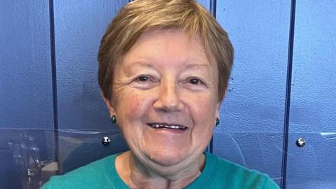 Woman with short light brown hair, round, black earrings and green sweater, head and shoulders shot. She is smiling at the camera