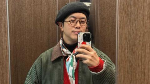 A man in a black beret and colourful scarf taking a photo in the mirror with his iPhone.