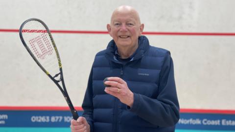 Martin Braybrook is stood in a squash court, positioned in the middle of the picture holding a squash racket in his right hard. He is holding a ball in his left hand close to his chest. He is smiling and looking towards the camera.