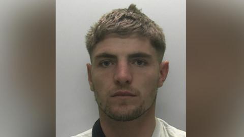 A young blond man with a white top looks at the camera.