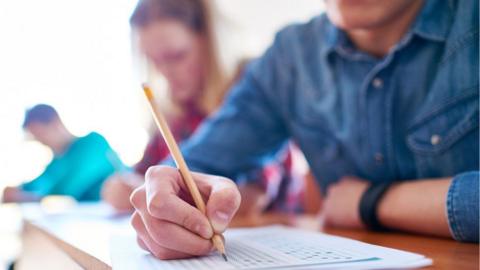 A student in non-school uniform holds a pencil and writes on a quiz sheet. Their face cannot be seen and other students behind him are blurred.
