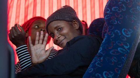 A woman in a grey hat sitting in a coach smiles and waves from the window at the people outside. 