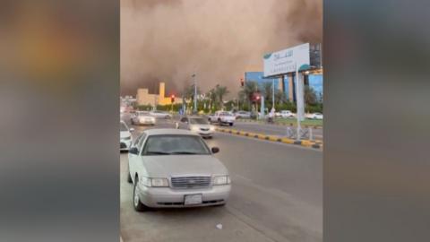 Cars drive down road as huge sandstorm cloud looms in the background 
