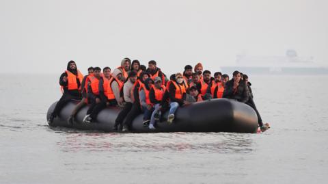 Migrants in orange life vests are seen packed onto a rubber dinghy as it moves across the sea.